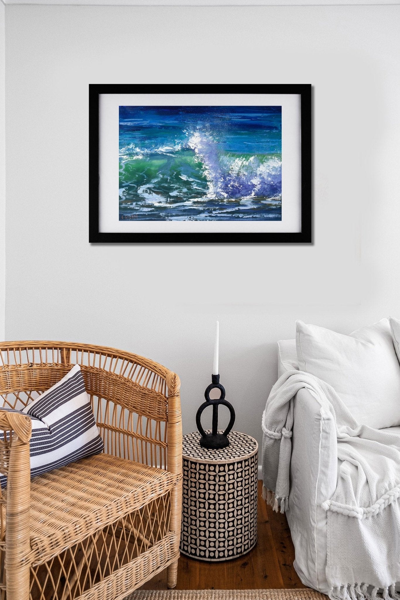 Framed artwork of a wave on a wall above a wicker chair and side table.