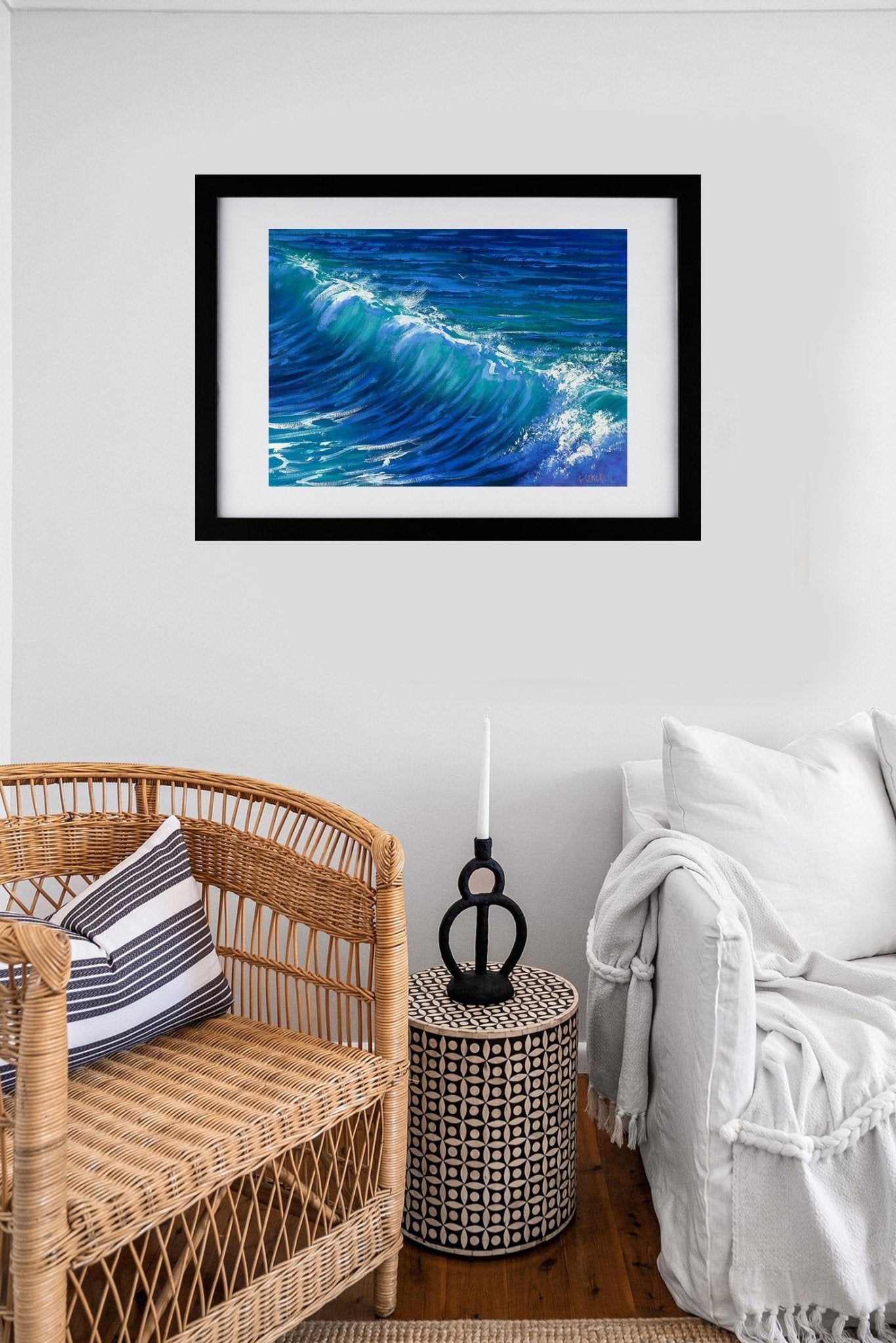Framed artwork of a blue wave on a wall above a wicker chair and side table.
