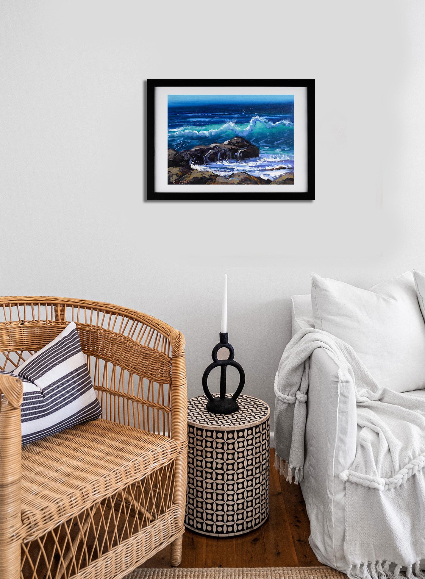 Framed artwork of a coastal scene above a wicker chair and side table in a room.