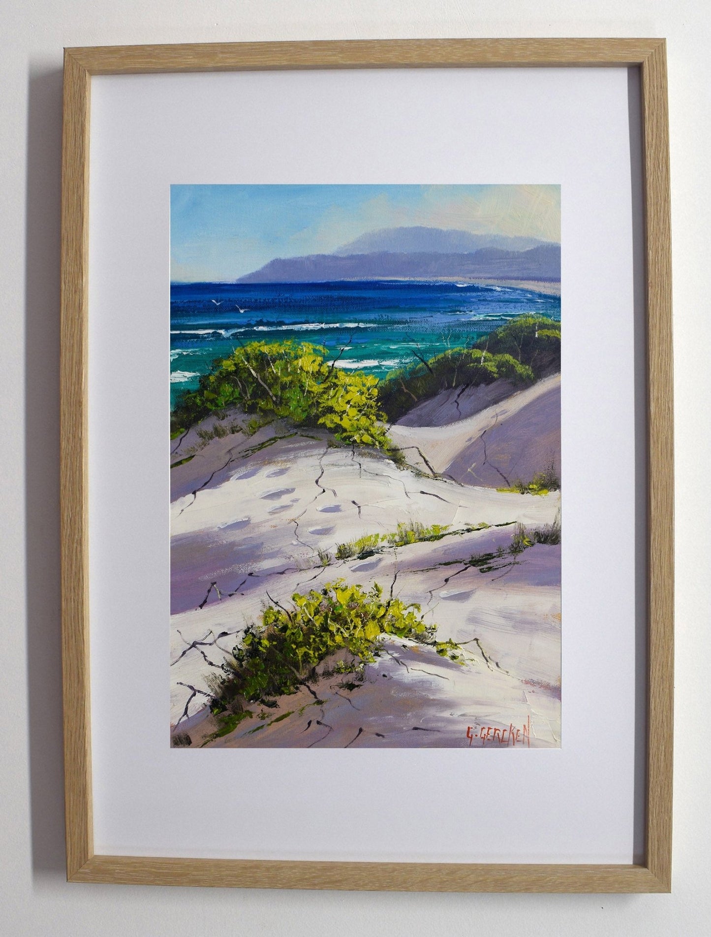 Framed painting of a coastal landscape with white sand, greenery, and blue water.