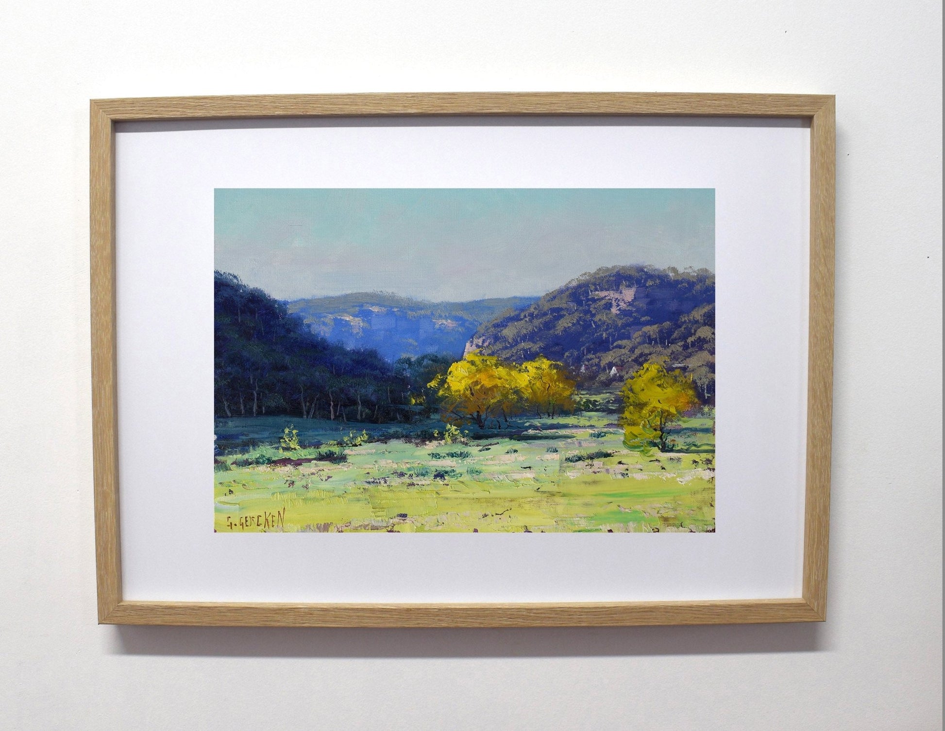 Framed landscape painting of a valley with trees and mountains on a white wall.